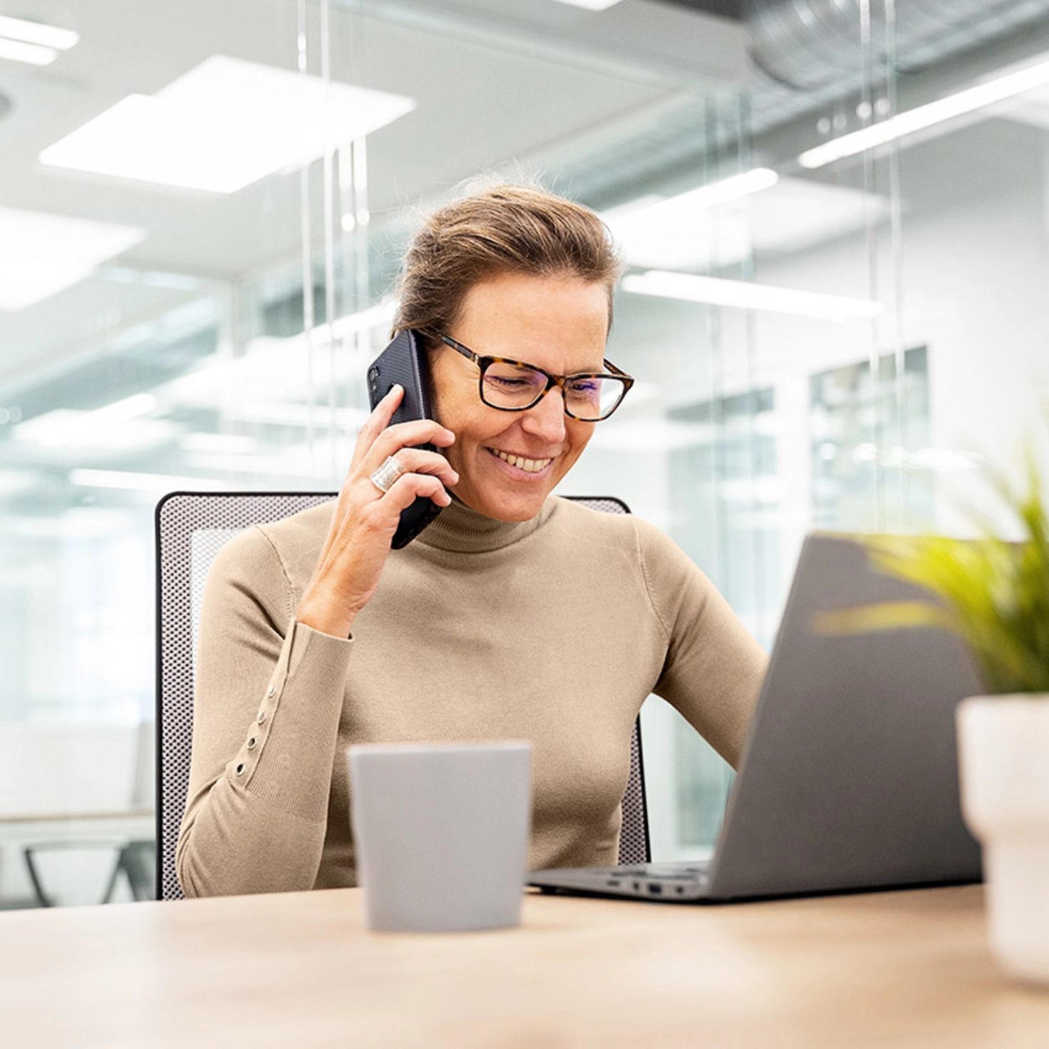 Une femme souriante est assise dans un bureau devant son ordinateur portable et téléphone avec son natel.