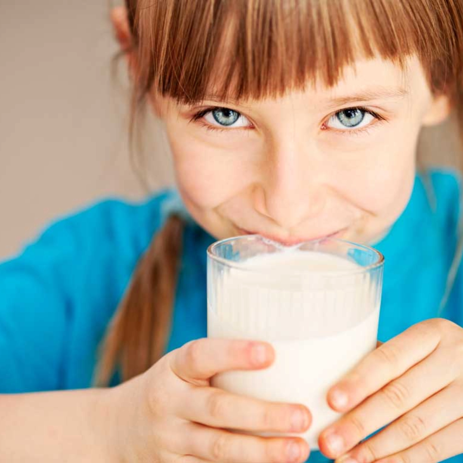 Ein Mädchen hält ein Glas Milch an seinen Mund und lächelt mit geschlossenen Lippen in die Kamera