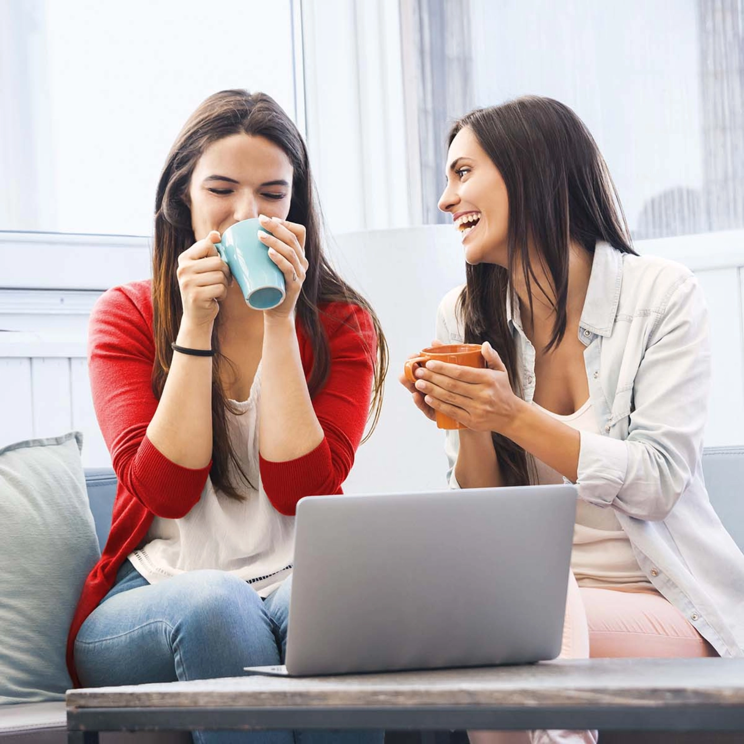 Two women are sitting on the sofa with a cup of coffee and talking. On the table in front of them is an open laptop.