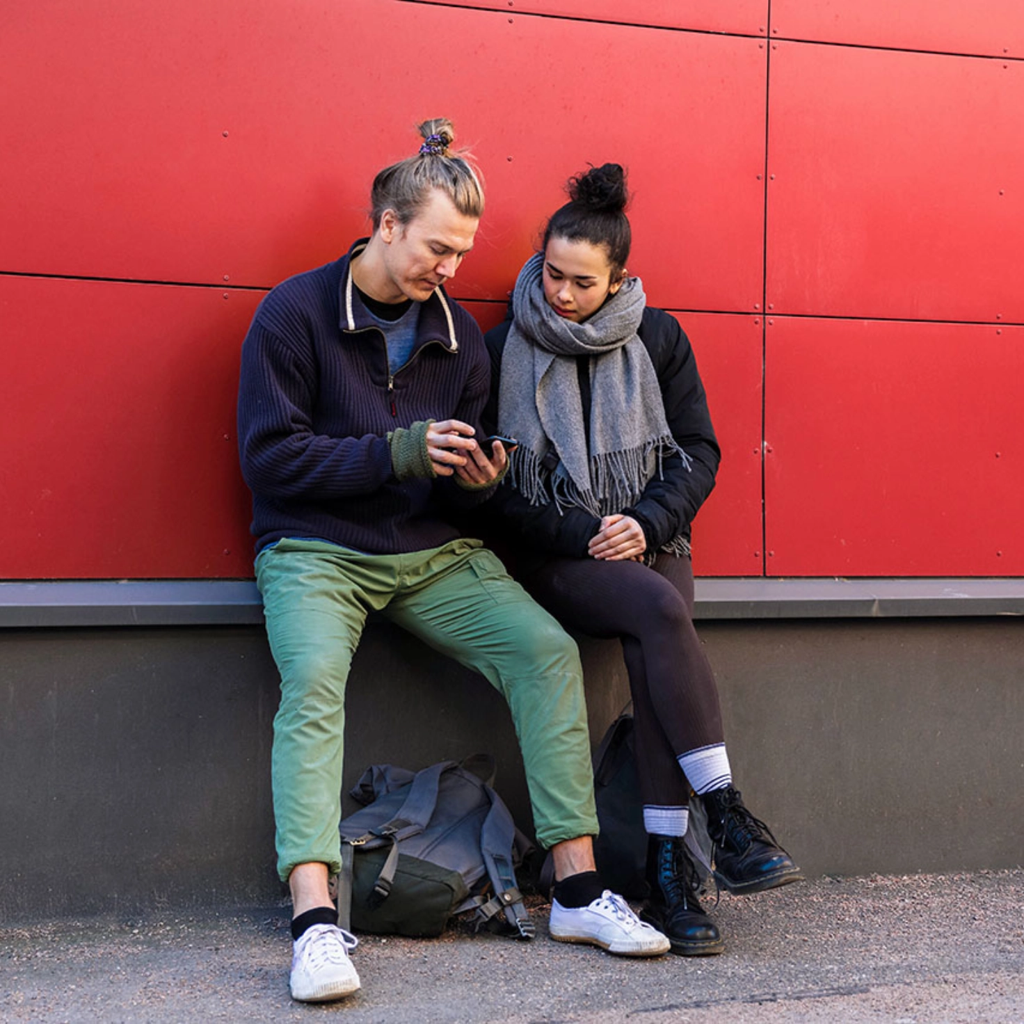 Un homme et une femme sont assis devant un bâtiment rouge et regardent ensemble son téléphone portable.
