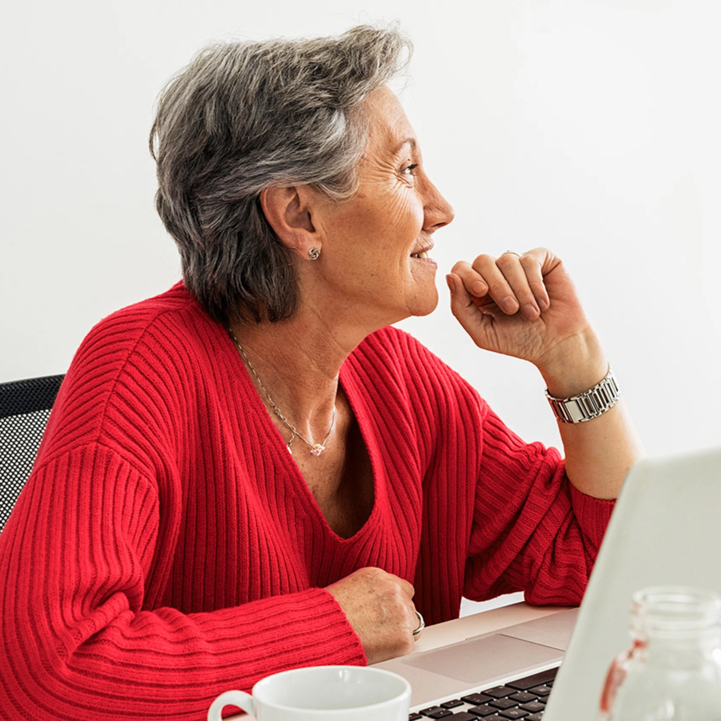 A woman with a friendly expression and a bright red jumper is sitting at the table with her laptop and thinking