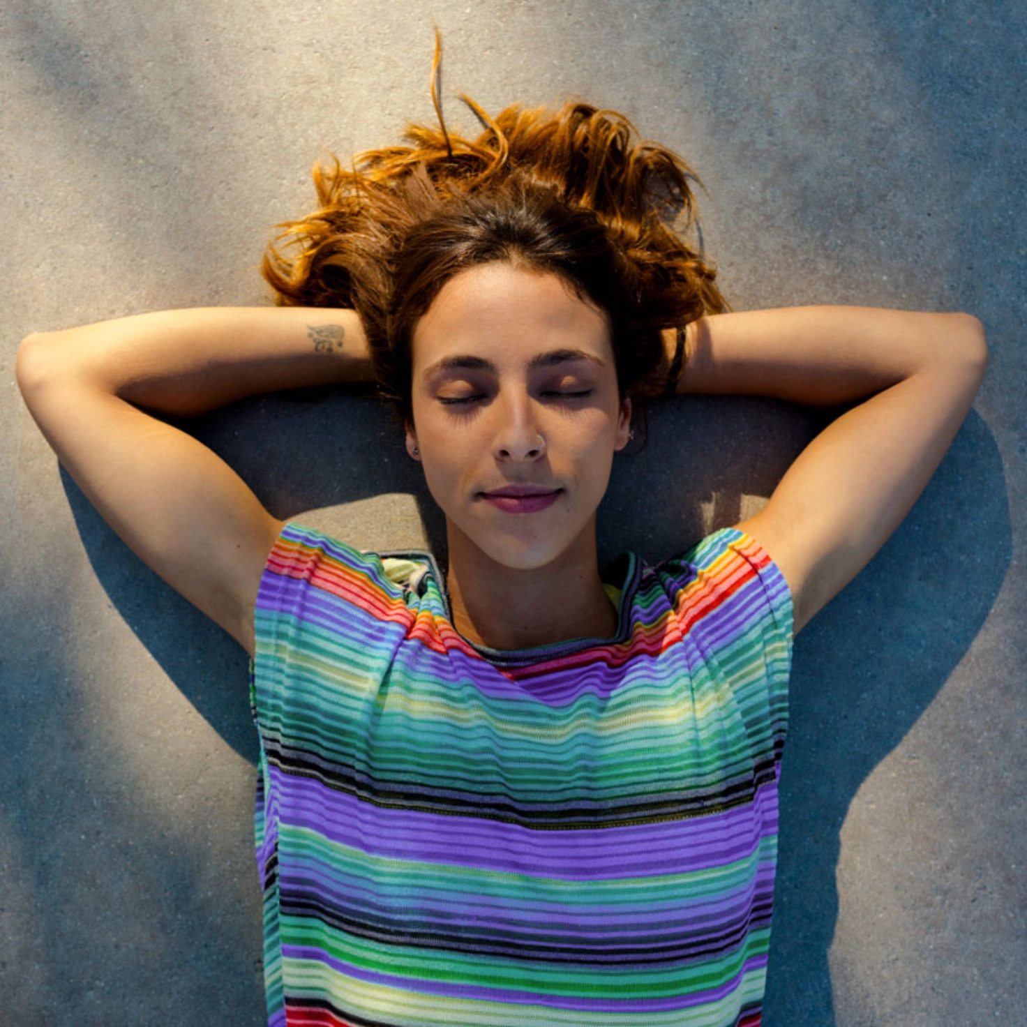 A young woman with a striped shirt lies on the ground with her eyes closed and enjoys the sun
