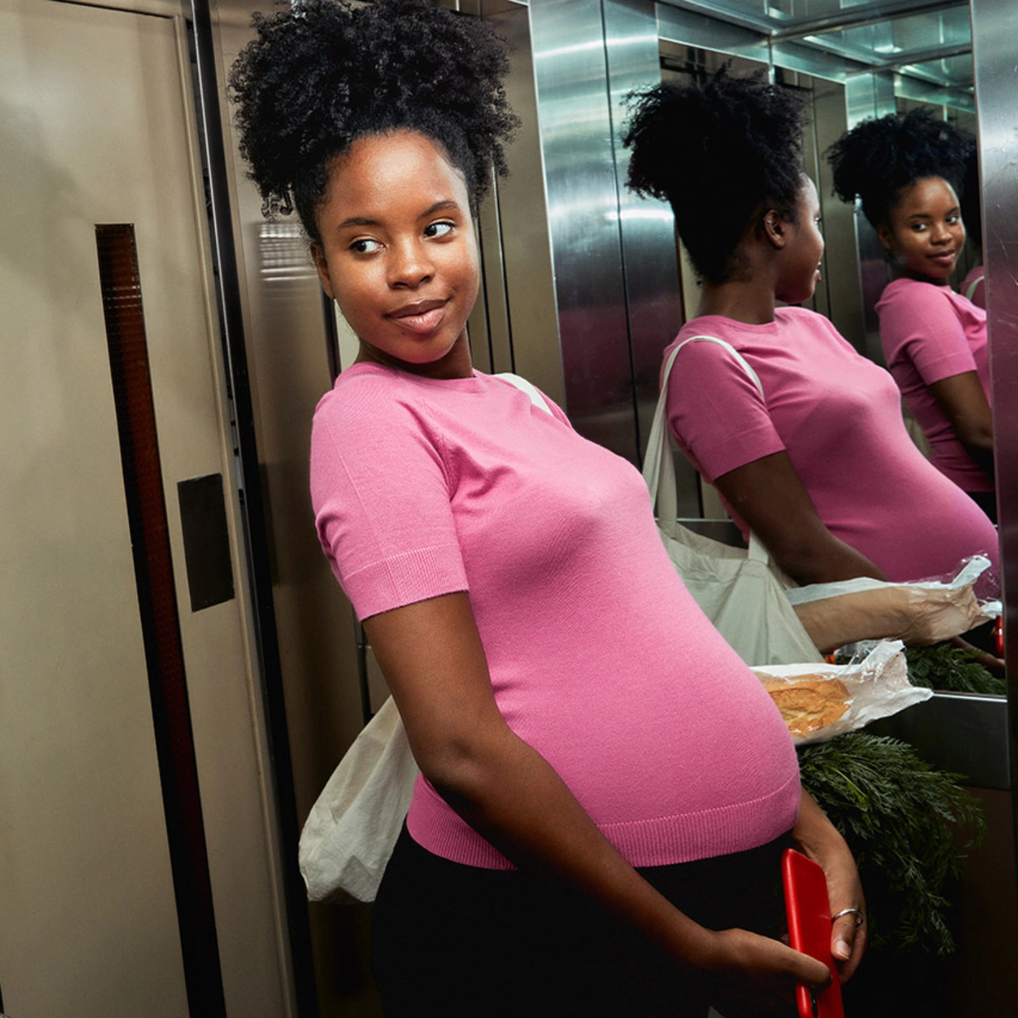 Une femme enceinte en chemise rose se tient dans l'ascenseur et regarde son ventre dans le miroir