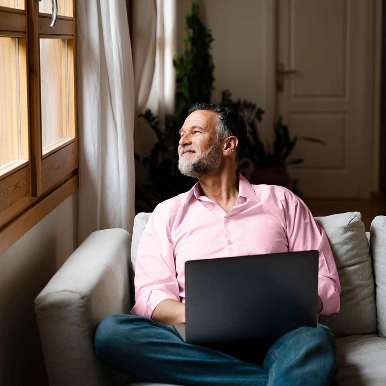 Man in a pink shirt sits on the couch with a laptop and looks out of the window