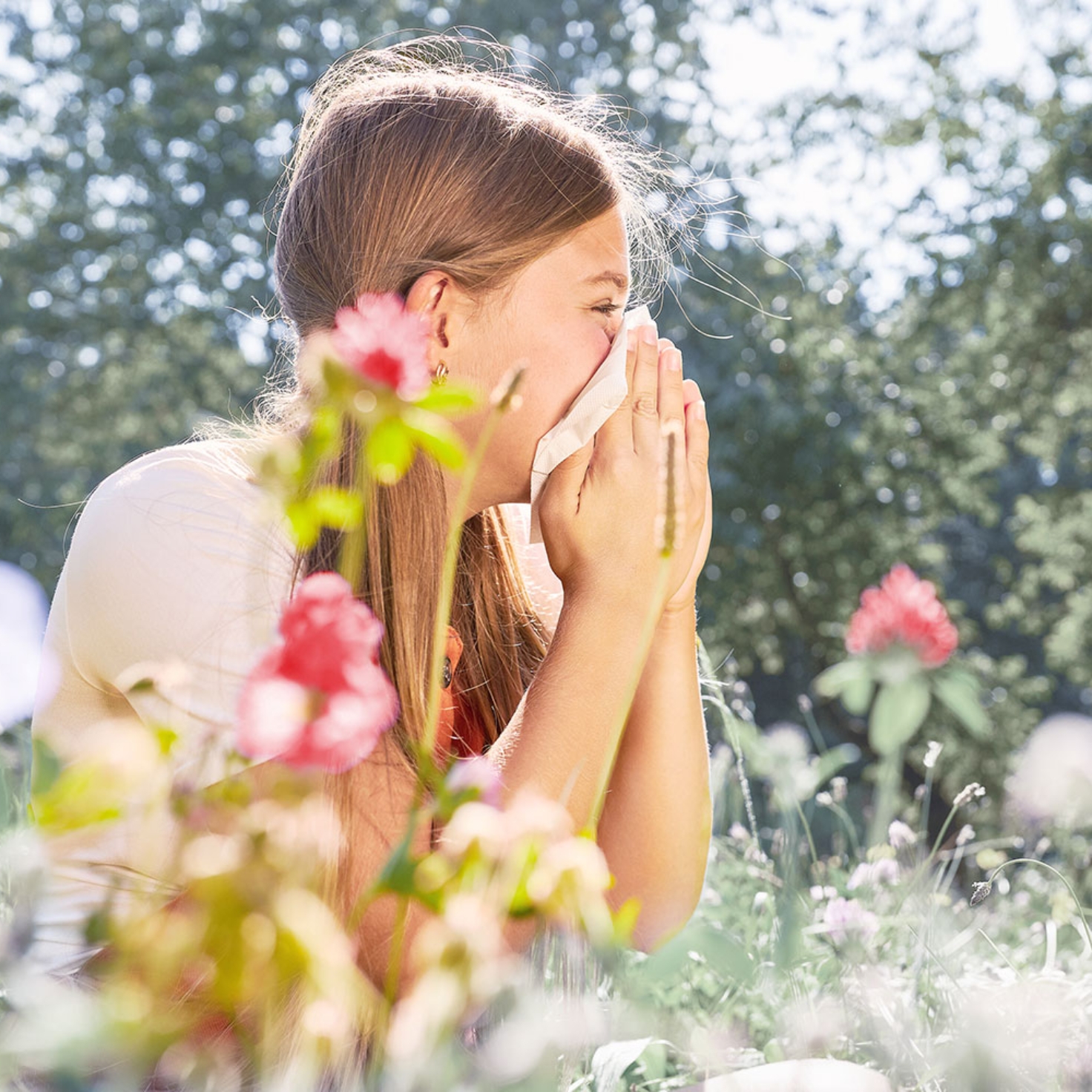 Ein Mädchen sitzt in einer Blumenwiese und niest in ein Taschentuch.