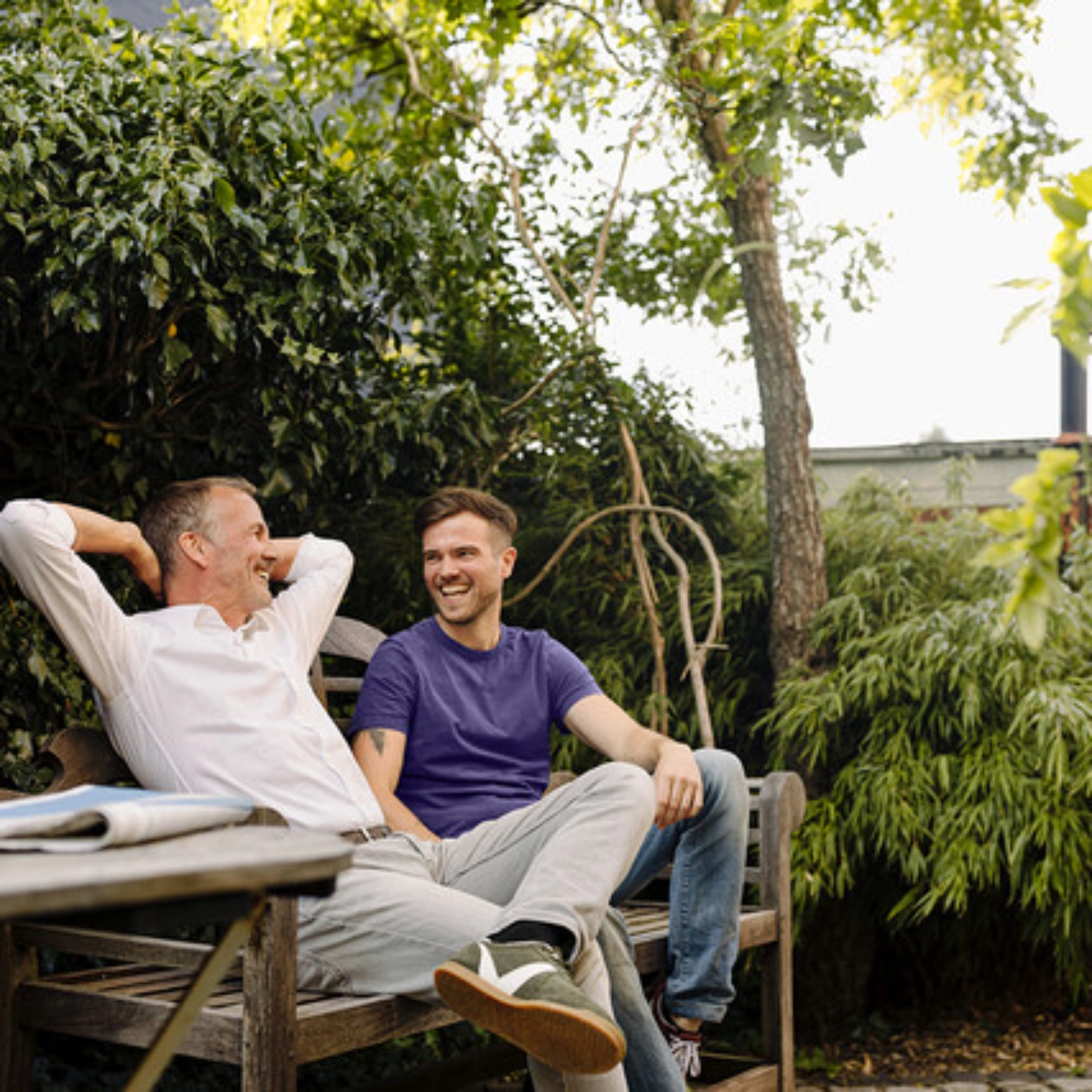 Ein Mann um die 50 und ein jüngerer Mann sitzen lachend zusammen auf einer Gartenbank. Im Vordergrund steht ein Gartentisch mit einer Tasse und einer Zeitung. Im Hintergrund befinden sich Sträucher und ein Baum.