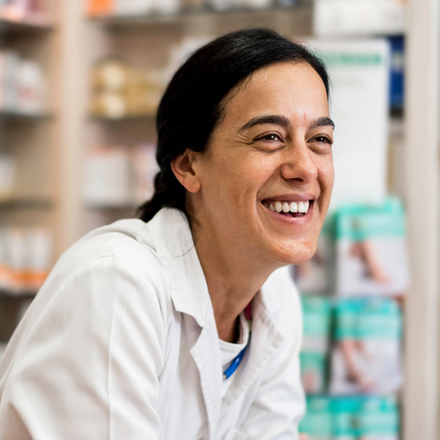 A pharmacist in a white coat laughs, behind her the shelves of the pharmacy can be vaguely recognised