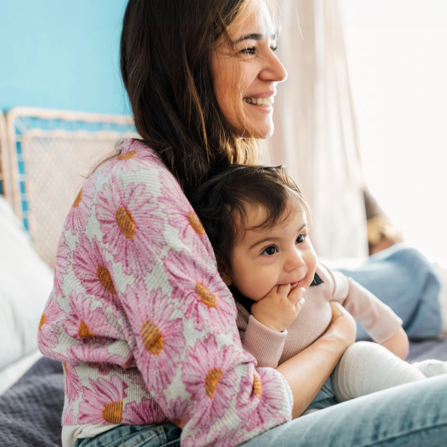 Une femme est assise dans son lit avec un bébé de quelques mois sur les genoux. La mère rit, le bébé met sa main dans sa bouche.