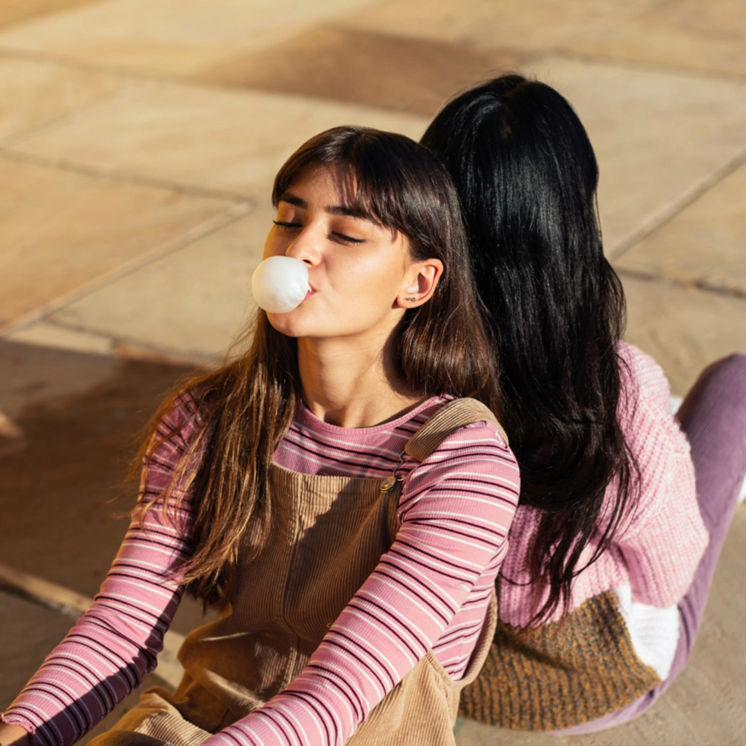 Two young women are sitting back to back on the floor, the one in front is making a bubble gum bubble