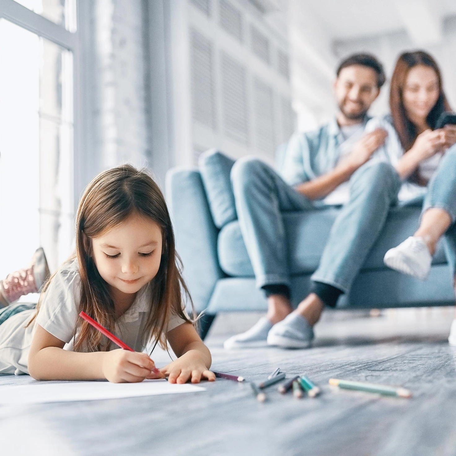 Mädchen liegt auf dem Boden und malt auf einem Blatt Papier während Eltern auf dem Sofa sitzen