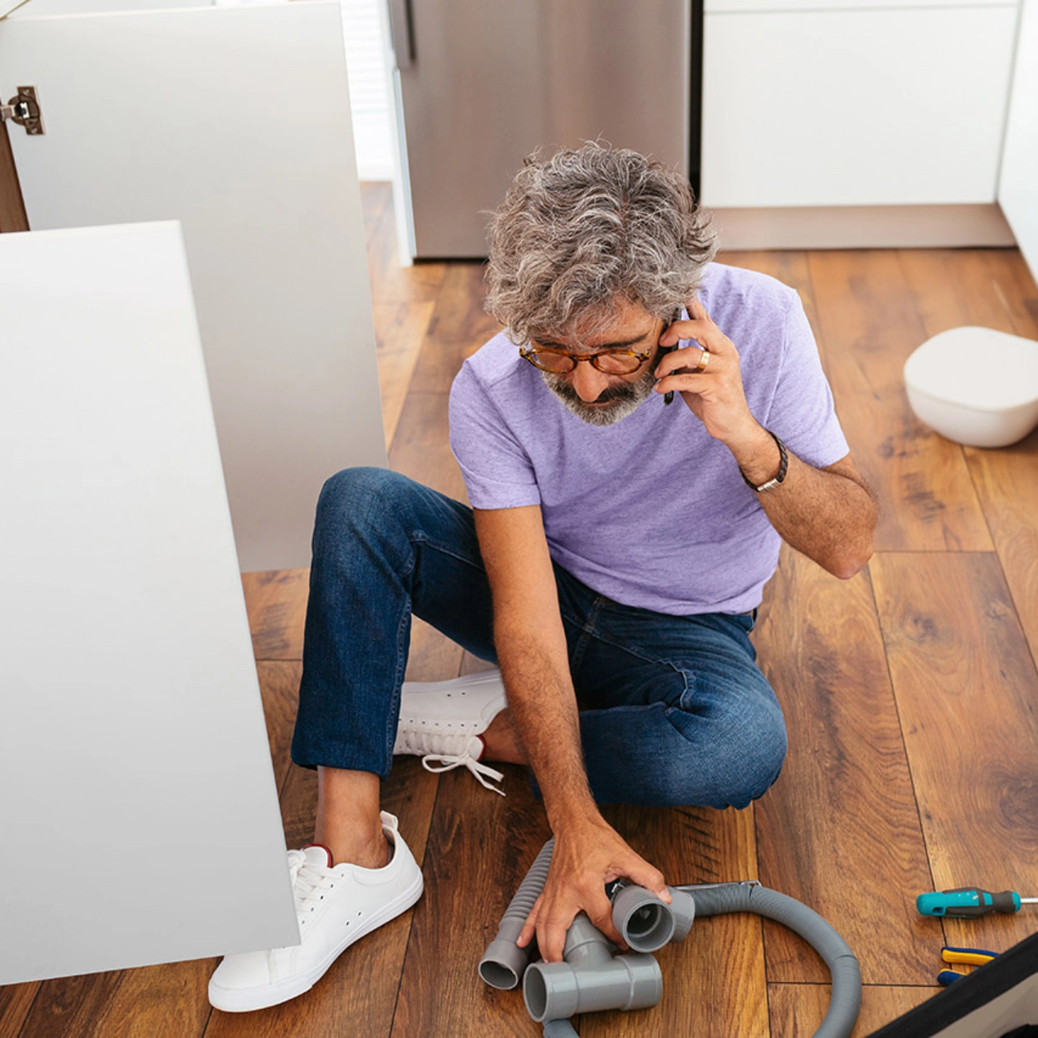 A man of about 50 is sitting on the kitchen floor, repairing something and talking on the phone
