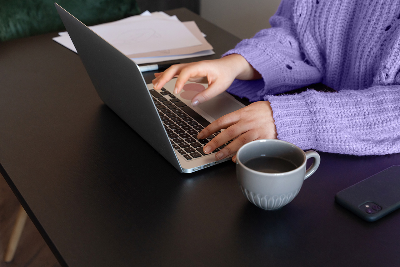 Una persona con un maglione viola sta scrivendo su un computer portatile con una tazza di caffè accanto a sé.