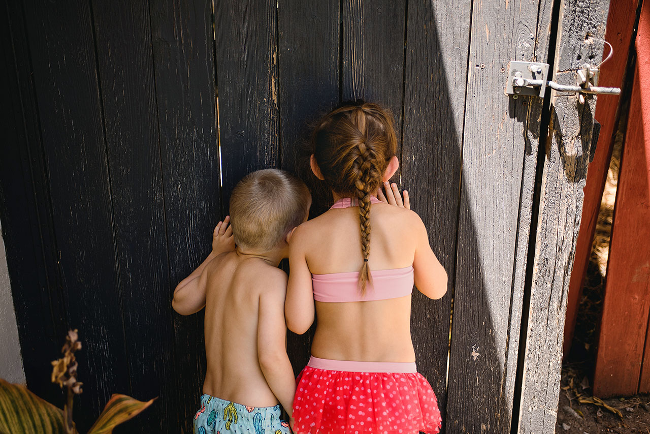 Un garçon et une fille en costume de bain regardent à travers une fente étroite dans une clôture en bois