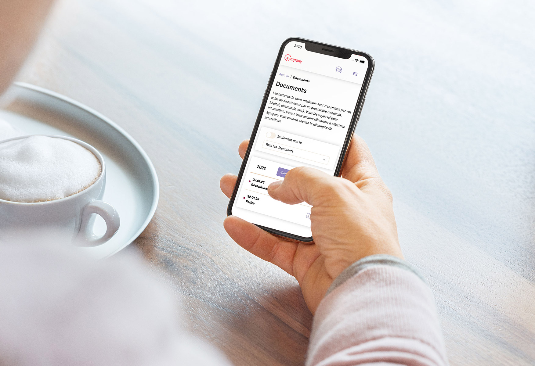 Une personne tient un smartphone dans la main. Sur l'écran, on voit le portail clientèle mySympany en version française.