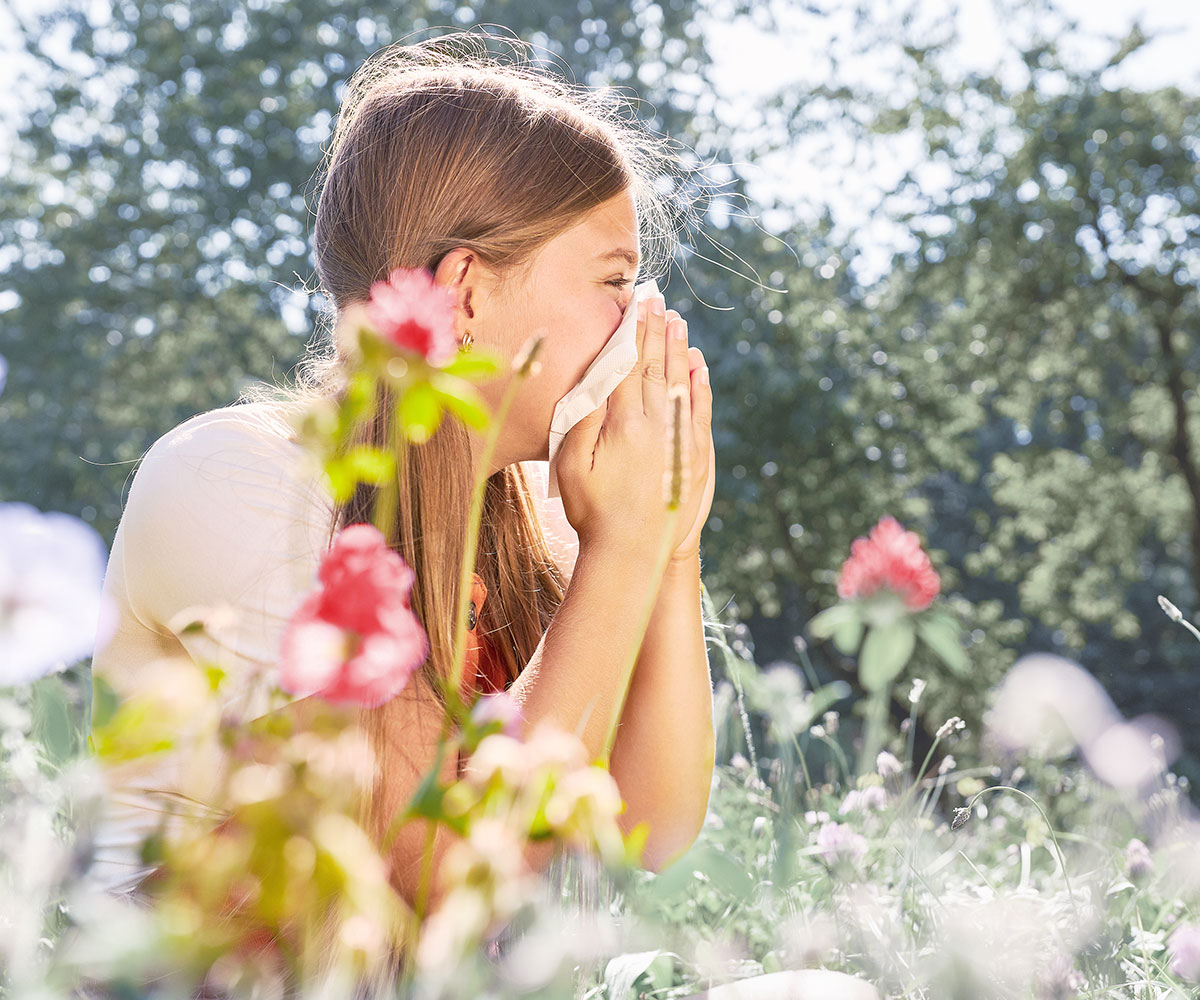 Ein Mädchen sitzt in einer Blumenwiese und niest in ein Taschentuch.