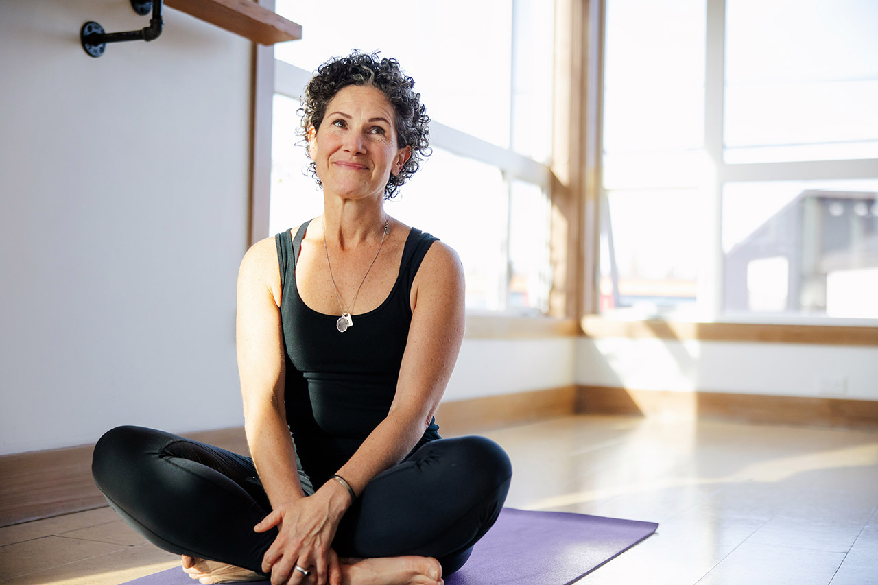 A woman with short, curly hair, a black shirt and black trousers sits cross-legged on a yoga mat