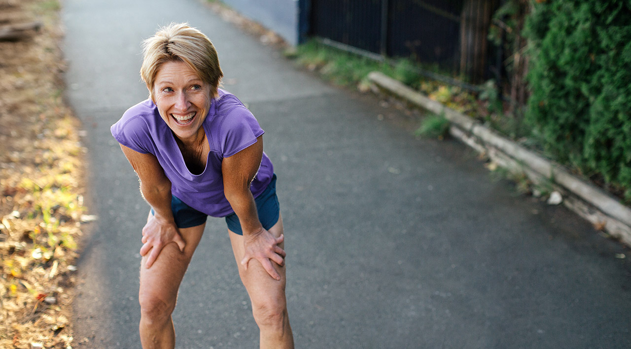 An older woman in short sportswear takes a break from jogging and leans on her thighs, laughing.
