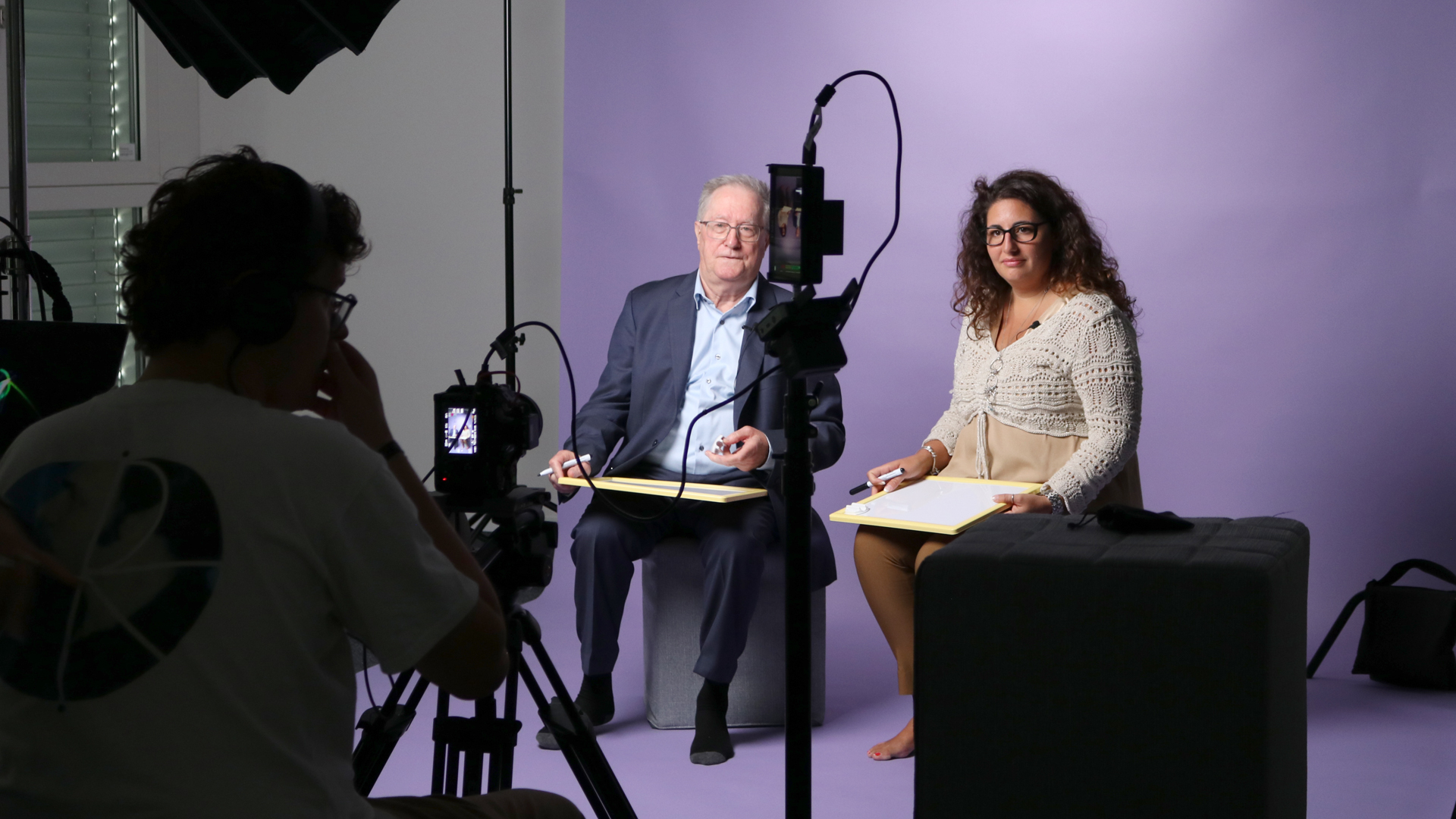Snapshot from the video shoot with the Sympany Customer Council: A man and a woman sit in front of a purple screen. The camera team can be seen from behind in the foreground.
