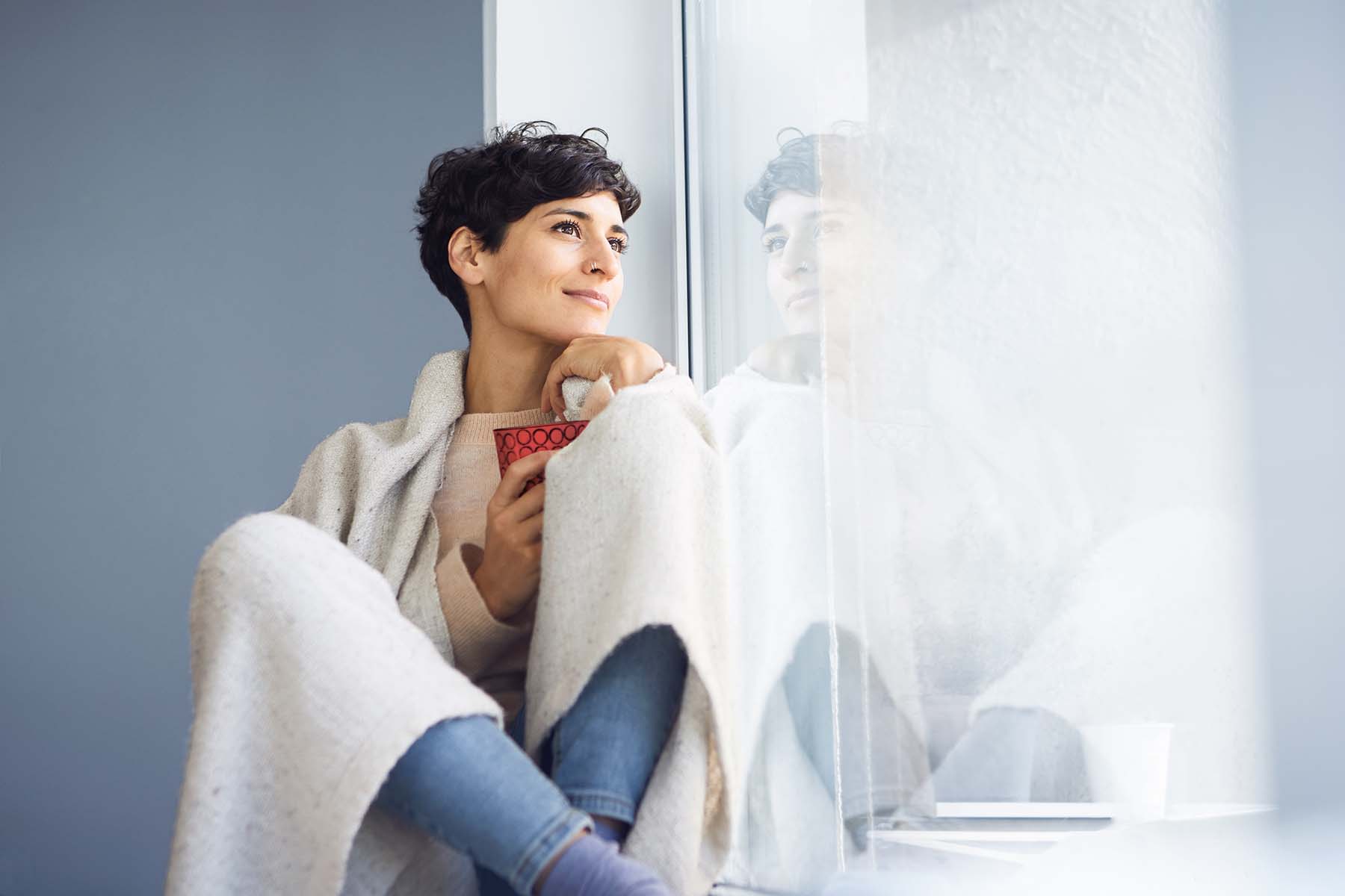 Frau sitzt mit Kaffee zu Hause am Fenster.