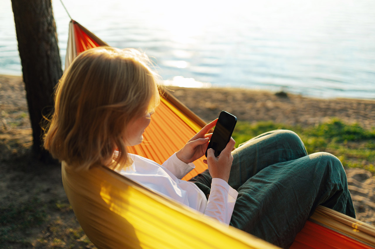 Eine Frau sitzt in einer gelben Hängematte an einem See und schaut auf ihr Handy