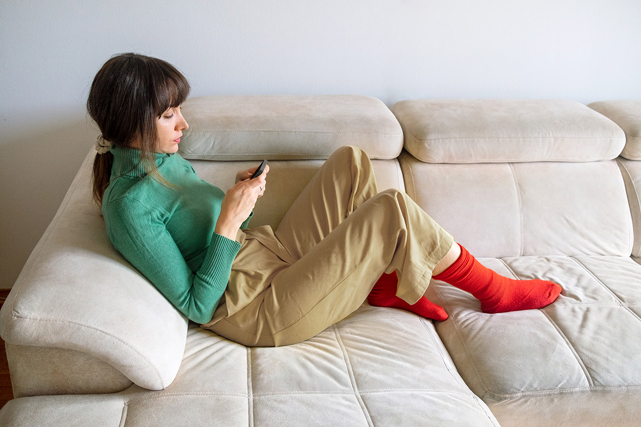 A young woman sits on the sofa with her legs bent, looking at her mobile phone