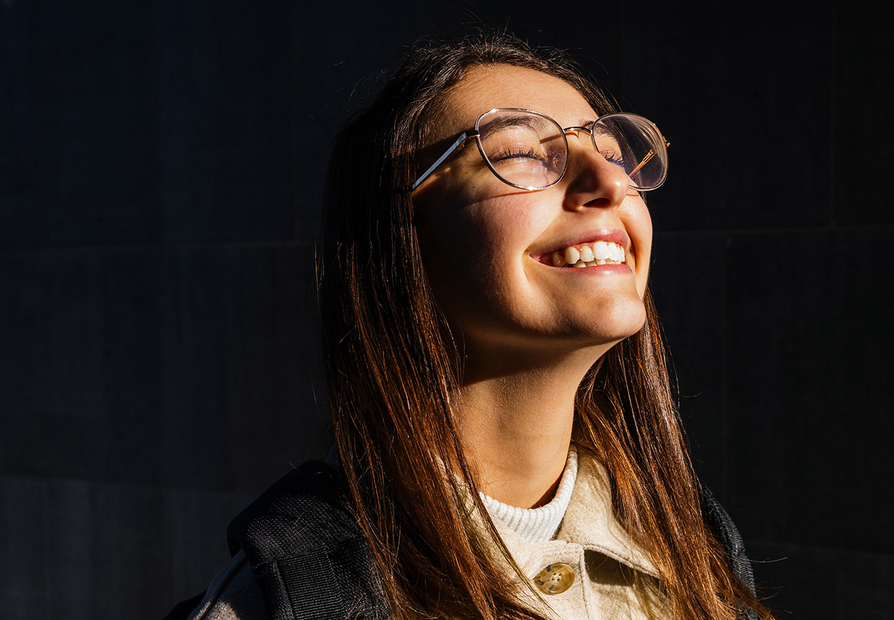 A young woman with glasses laughs with her eyes closed, the sun shining on her face